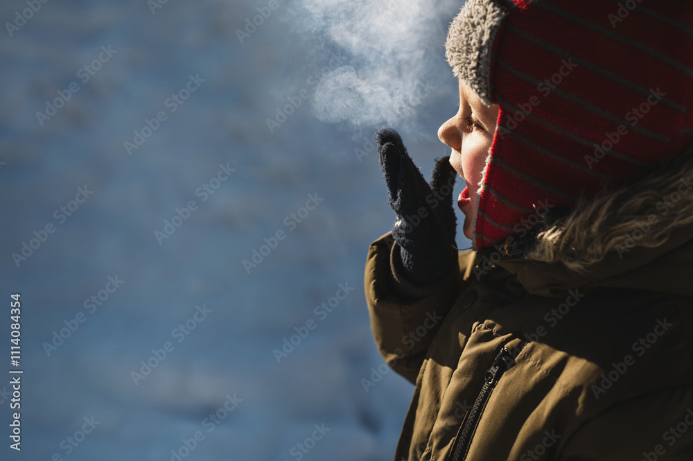 Fototapeta premium 4 year old boy in winter hat and jacket inhales frosty air and exhales steam. breathe out steam outdoor in winter