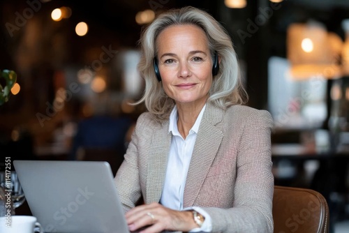 A mature professional woman focuses on her laptop in a vibrant cafe, wearing headphones, absorbed in work, representing modern remote work culture and digital lifestyle. photo