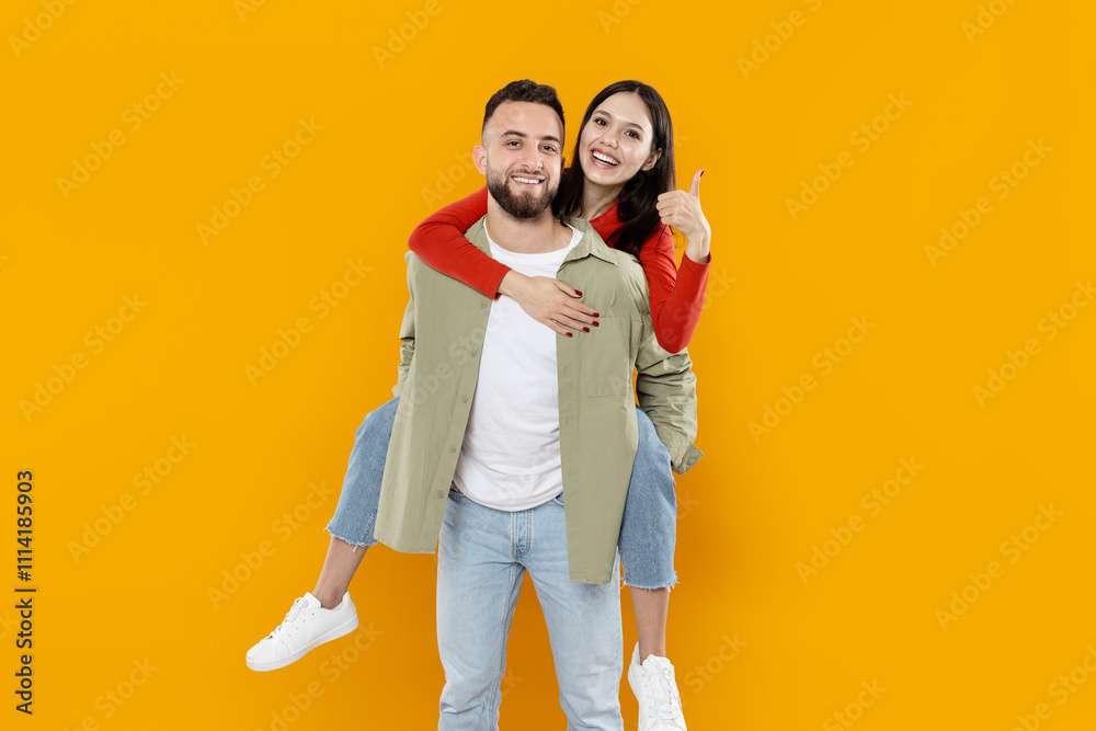 A joyful couple shares a fun moment, with the woman giving a thumbs up while riding piggyback on her partner. The bright yellow backdrop adds to the cheerful atmosphere of their playful interaction. A joyful couple shares a fun moment, with the woman giving a thumbs up while riding piggyback on her partner. The bright yellow backdrop adds to the cheerful atmosphere of their playful interaction.