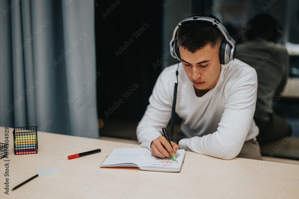 A young student wearing headphones intensely focused on writing notes in a notebook at a desk in a classroom. A young student wearing headphones intensely focused on writing notes in a notebook at a desk in a classroom.