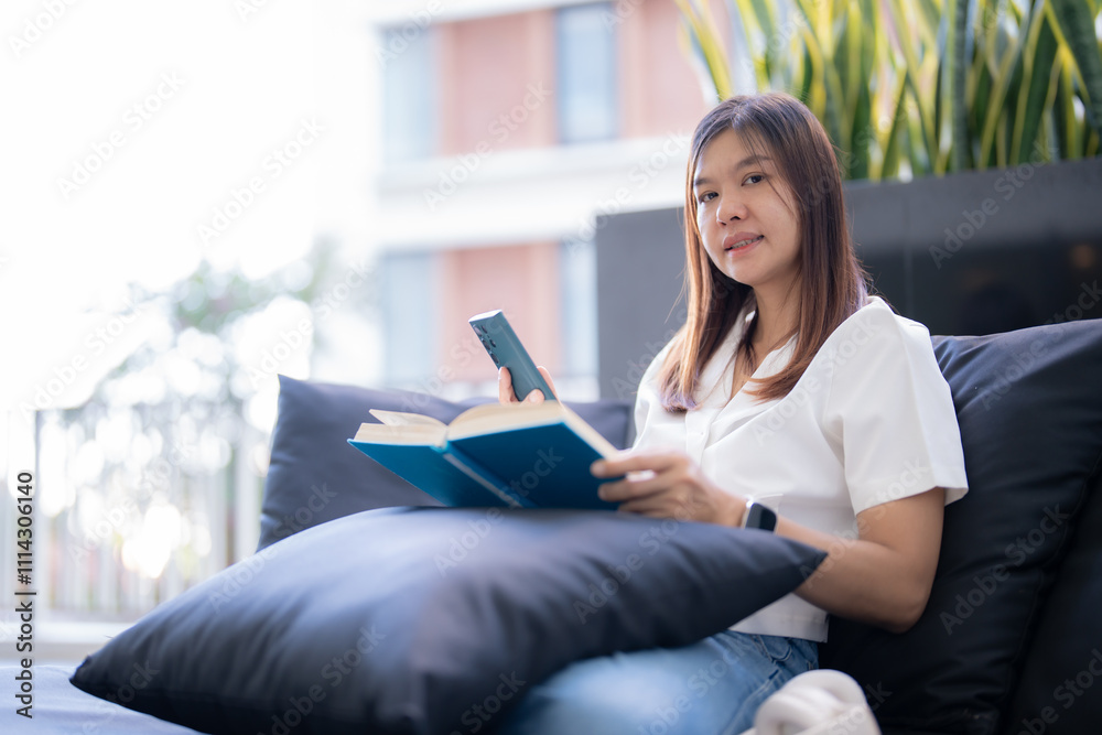 Young asian woman is sitting comfortably on a modern sofa, enjoying a moment of leisure by reading a book while holding her smartphone, possibly taking a break from work or studies Young asian woman is sitting comfortably on a modern sofa, enjoying a moment of leisure by reading a book while holding her smartphone, possibly taking a break from work or studies