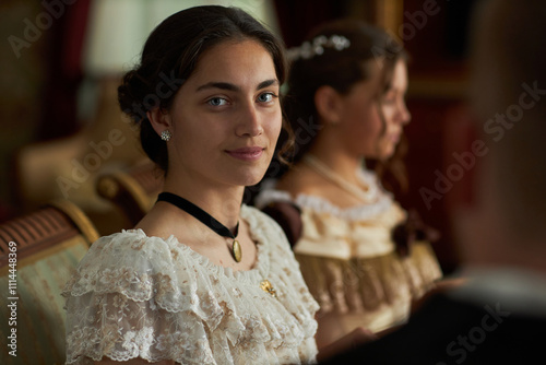 Portrait of young woman wearing lace gown gazing softly at camera with another lady in background in classic interior photo