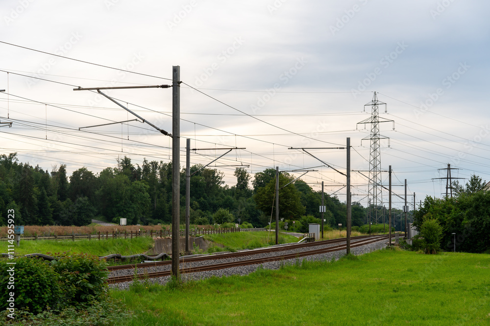 Fototapeta premium Infrastructure of railroad and energy supply come together in this photograph: High voltage pylons of different sizes, and the railroad tracks and overhead contacts. Street besides railroad tracks.