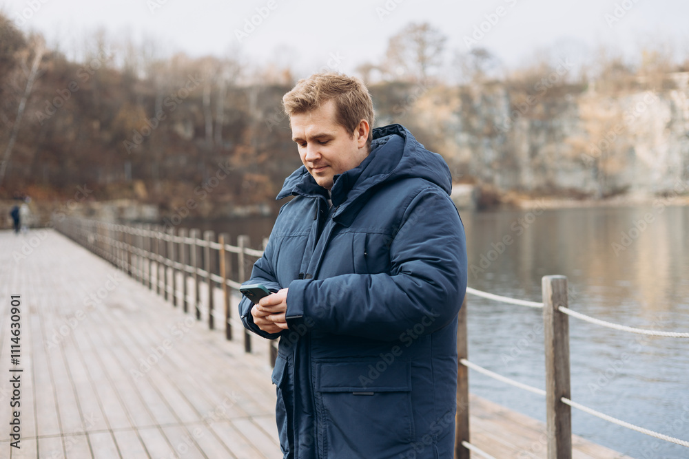 Adult 30s man hiker stand outdoors in a natural setting, smile while using his mobile phone outdoors. Happy man standing on the lake wooden pier.