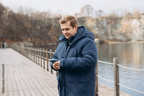 Obraz na płótnie Adult 30s man hiker stand outdoors in a natural setting, smile while using his mobile phone outdoors. Happy man standing on the lake wooden pier. w salonie