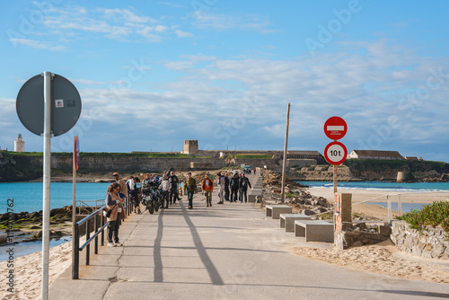 People walk along a path towards the historic Castle of Guzman in Tarifa, Spain. Sandy beaches, clear waters, and a lighthouse enhance the coastal scene. photo