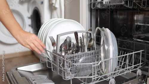 Female hand removing clean cutlery from a loaded dishwasher. Background of open wash machine 