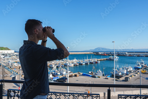 A man uses binoculars on a balcony overlooking a bustling marina with numerous boats, set against a clear blue sky and distant mountains. photo