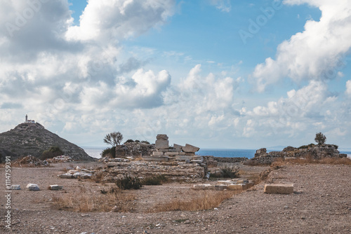 An ancient archaeological site near the coast, featuring scattered ruins with a serene sea and a lighthouse visible on a hill in the background under a partly cloudy sky.