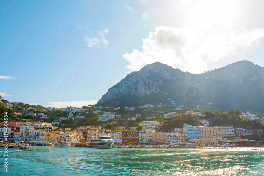 The photo captures a view of Capri from the water, showcasing the island's iconic cliffs, colorful buildings, and lush greenery set against the clear blue sea and sky.