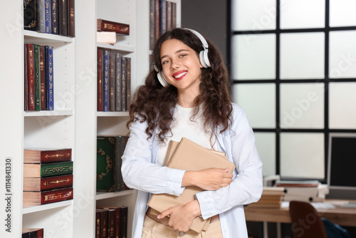 Smiling young woman with headphones holding books in library, education and l...