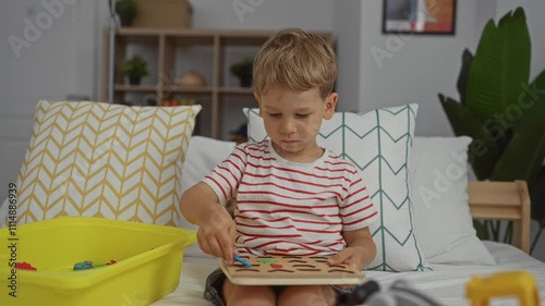 Cute blonde toddler boy playing with a wooden puzzle on a comfortable bed in a cozy bedroom with bright pillows and shelves in the background.