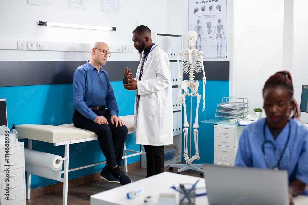 Obraz premium Black doctor offers health advice to senior white man during medical checkup holding bottle of prescribed pills. Pensioner patient receives friendly consultation on healthcare plan and medication.