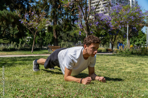 A young man with brown hair holds a plank position outdoors in a park, concentrating on his workout
