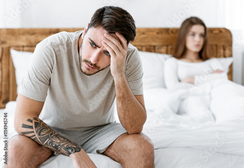 Problem in bed. Man sits on bed and holds on his head, woman is offended and crossed her arms