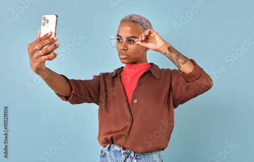 Confident African American individual poses against pastel blue backdrop, taking a stylish selfie. With vibrant attire and playful expression, they showcase their unique fashion sense and personality