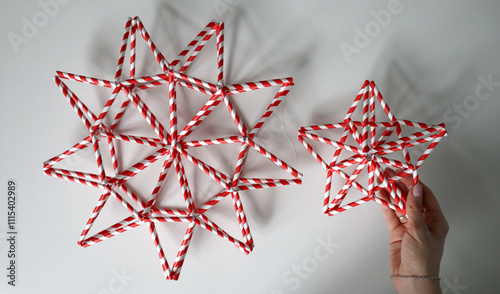 Female hand holds star. Nearby is larger star of different shape. Stars from tubes in red and white stripe. White threads are visible. They cast shadow. White background. Idea for decoration. Holiday. photo