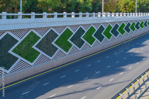 The side direction signs on eight-lane highway are saddled with green artificial plants. On the asphalt there is discontinuous white and yellow solid strip, dividing metal bumpers, no cars, no people