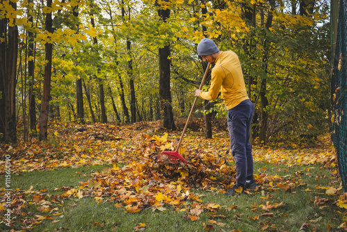 Man in a yellow sweater and beanie raking autumn leaves in a colorful forest. The golden foliage and serene atmosphere showcase the beauty of seasonal outdoor work
