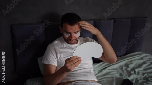 30-year-old man with a silk mask for sleep applying patches under the eyes from swelling, wrinkles,