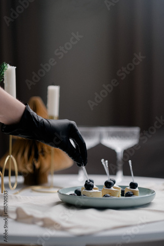 In the image, a gloved hand is delicately placing a blueberry on a piece of cheese. 