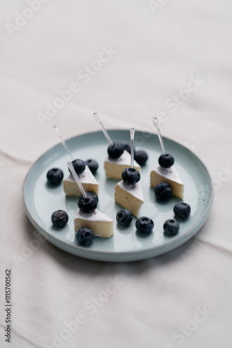 In the image, a gloved hand is delicately placing a blueberry on a piece of cheese. 