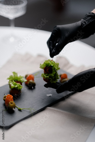 In the image, a gloved hand is gently arranging a small salad appetizer on a black slate plate. 