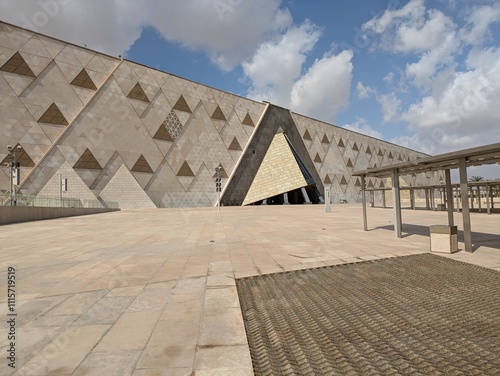 New Grand Egyptian Museum, mitte der Fassade befindet sich ein markant eingeschnittener, dunkler Dreieckseingang des Museums mit heller Frontfläche, darüber ein Himmel mit einigen weißen Wolken.