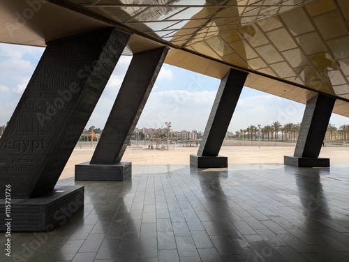 New Grand Egyptian Museum stark schräg stehenden, massiven Stützen zu sehen, der golden schimmernde Eingang zum Museum, Palmen und vereinzelten Strukturen in der Ferne unter leicht bewölktem Himmel.