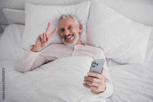 Relaxed mature man enjoying morning in bed with smartphone and peace gesture photo