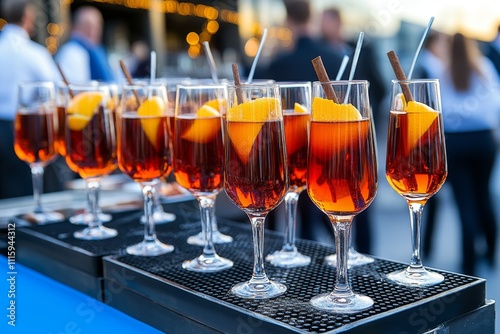  Copas transparentes llenas de vino especiado decoradas con rodajas de naranja y ramas de canela, servidas al aire libre durante un evento.
 photo