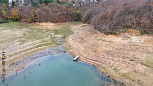 Dried-up Reservoir Reveals Exposed Shoreline and a Small Boat. High-angle, full shot of a reservoir that has significantly decreased in water level, revealing exposed, layered earth and barren shoreli photo