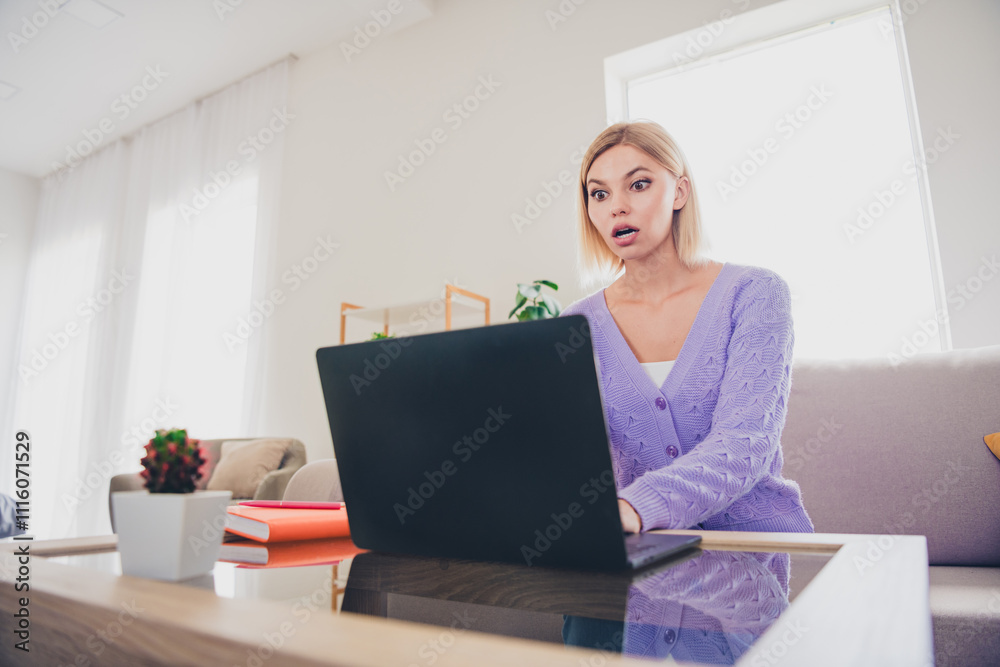 Fototapeta premium Young woman wearing a knitted sweater looking surprised at laptop in bright living room during daytime