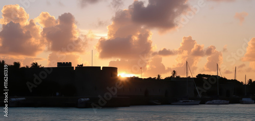 An atmospheric, sunset-lit depiction of Fort Charlotte in Nassau, with its imposing stone walls and cannons dramatically silhouetted against a vibrant turquoise harbor. photo