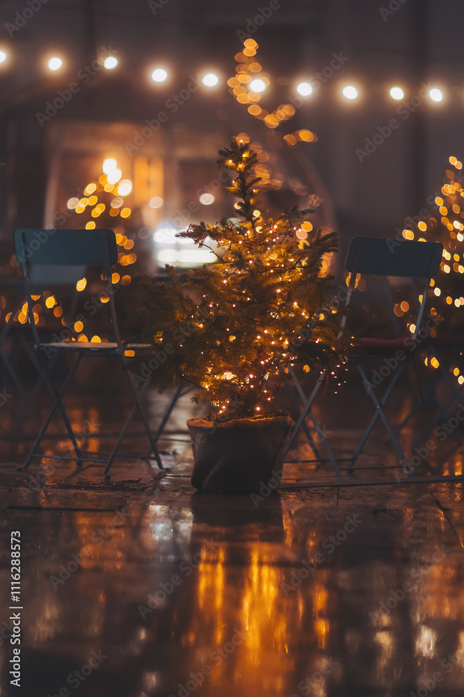 Obraz premium Christmas tree in pot on city street near cafe. Restaurant terrace decorating potted xmas trees. Focus on the Christmas tree in the foreground. lights bokeh.