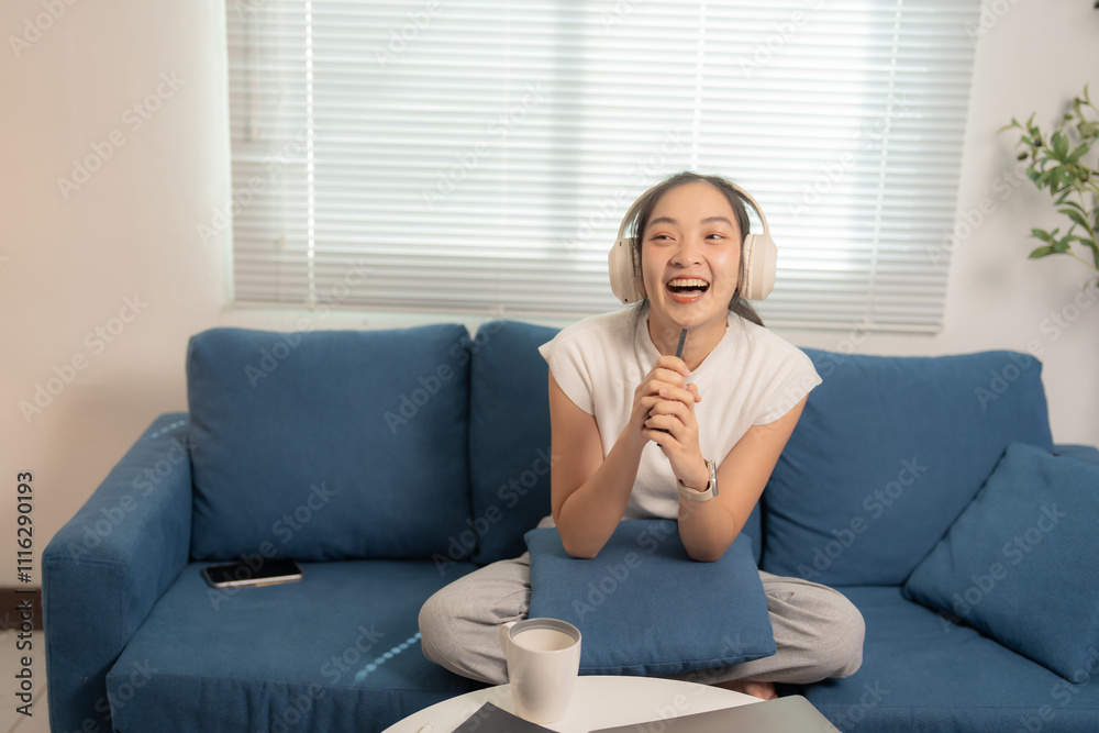 Happy freelance woman wearing headphones, singing and holding a pen like a microphone while relaxing on a comfortable sofa with a laptop and coffee nearby Happy freelance woman wearing headphones, singing and holding a pen like a microphone while relaxing on a comfortable sofa with a laptop and coffee nearby