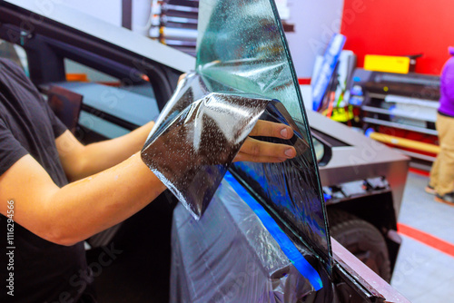 Worker technician applies window tint film to vehicle in workshop setting, focusing on precision quality.