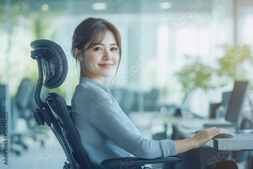 Young Asian Businesswoman Smiling in Modern Office Environment, Seated in Ergonomic Chair, Working on Computer, Professional and Relaxed Atmosphere, Natural Light Filling Space photo