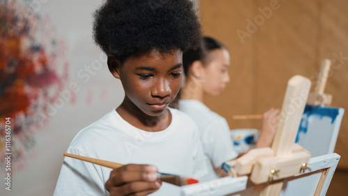 Quadro su tela African boy painting canvas with watercolor while happy caucasian girl draw cool tone picture at colorful stained wall