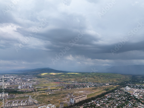 Mountain landscape in summer before the rain. The sun rays break through the clouds. City near the mountains. Top view, drone photo.