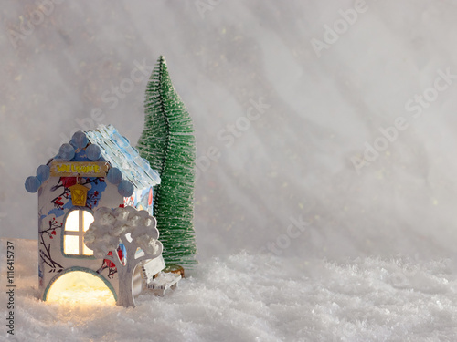 A toy house with a Christmas tree behind it stands in the snow. New Year background