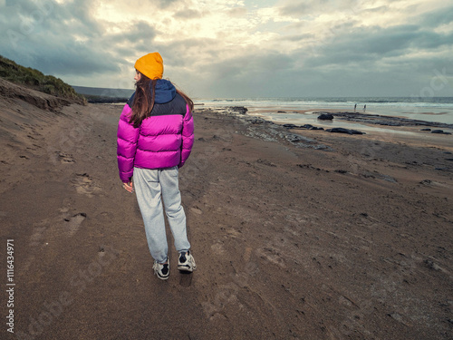 Teenager girl walking on stunning Fanore beach in county Clare, Ireland. Travel and tourism concept. Blue ocean, dramatic sky and sun. Warm sunny day. Model wears purple jacket and grey pants. photo