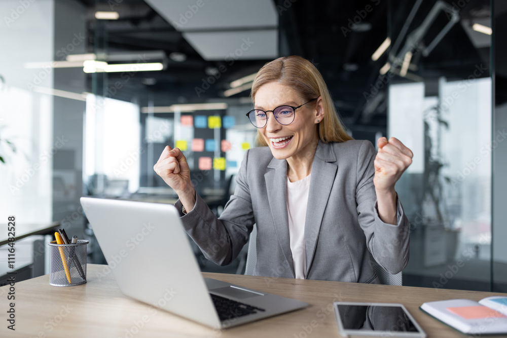 Fototapeta premium Mature businesswoman expressing excitement as she celebrates a work success. The image captures her joy and enthusiasm in a modern office setting while working on her laptop.