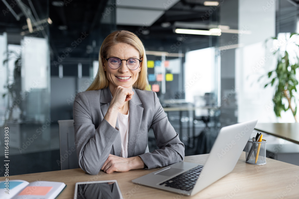 Fototapeta premium Mature woman businesswoman smiling confidently while working on a laptop in a modern office. Her professional demeanor and stylish attire reflect success and leadership qualities.