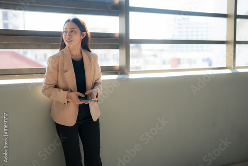 Young asian businesswoman wearing a beige blazer and black pants is using her smartphone while standing next to a large window in a modern office building, looking away pensively photo