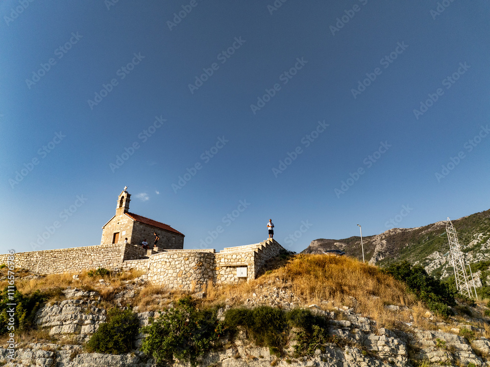 Young Man Standing on the Edge of a Fence at Sveti Stefan Church with Mountains in the Background
