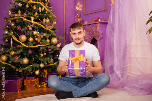 young man in white t-shirt and jeans on purple christmas background holding gift box . christmas tree with toys