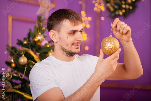 young man in white t-shirt and jeans on purple christmas background holding christmas tree toy in hands . christmas tree with toys