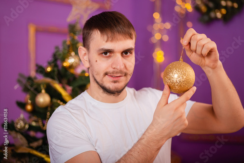 young man in white t-shirt and jeans on purple christmas background holding christmas tree toy in hands . christmas tree with toys