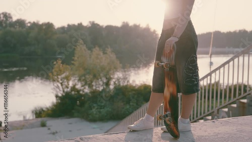 Teen Skater Girl Silhouette at Sunset | Slow-Mo Skateboard Stance by River | Backlit Urban Scene with Blurred Railings | Teenager's Board Vertical Grip | Golden Hour Skate Culture Snapshot photo